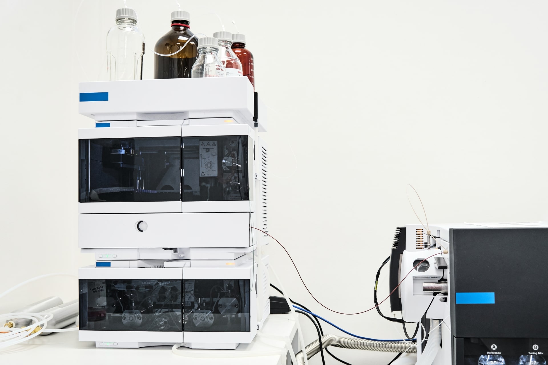 A stack of refurbished LC-MS and GC-MS equipment with glass bottles on top, connected by tubes to a black device with a blue stripe.